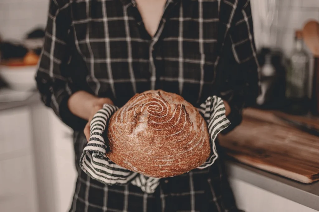 Ženska sredi kuhinje v rokah drži sveže pečen domač hlebec kruha.