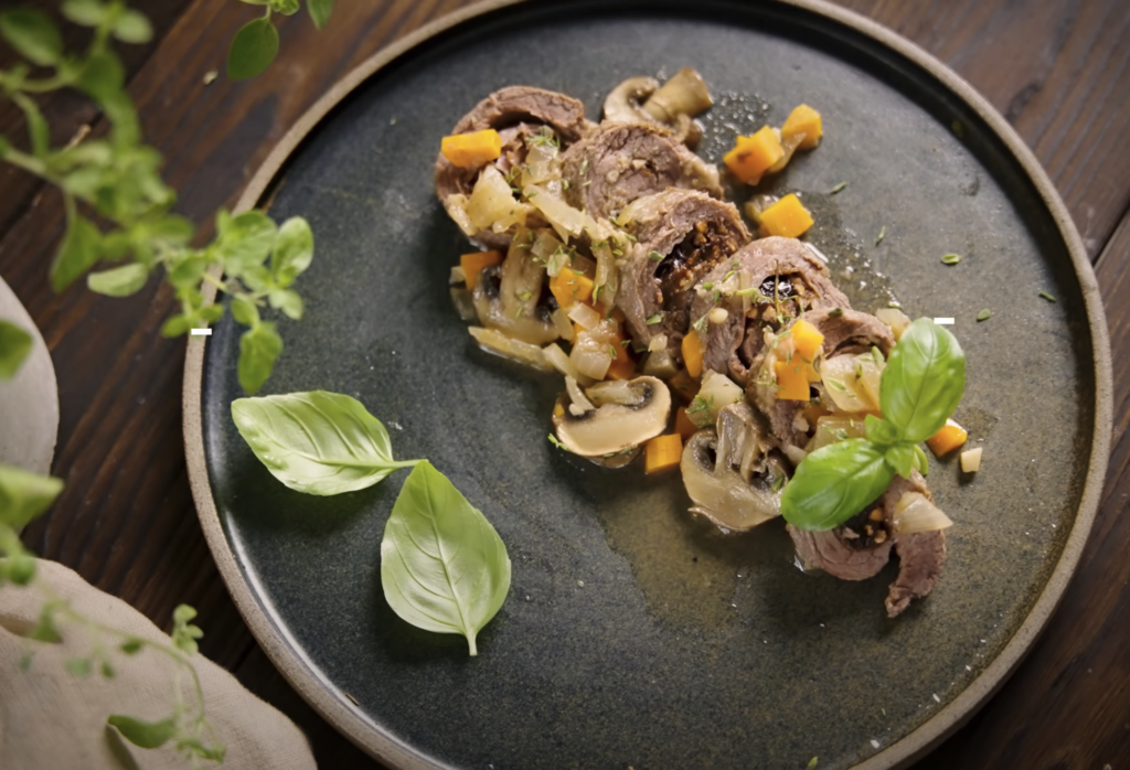 Na sliki je črn krožnik, na keterm so razporejeni pečeni goveji zvitki s slivami.